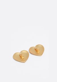 Un par de pendientes de clip en forma de corazón en tono dorado con un acabado suave y el nombre de la marca grabado en la parte posterior, exhibidos sobre un fondo blanco.