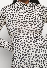 Långärmad polotröja i ett lätt och elastiskt material med en vit bas och svarta cirkulära mönster i varierande storlekar.