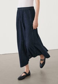 Jeanskjol i mörkblått med en flödande design, med synliga sömdetaljer. Bärs med svarta platåskor, vilket framhäver tygtexturen.