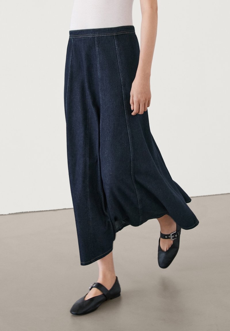 Jeanskjol i mörkblått med en flödande design, med synliga sömdetaljer. Bärs med svarta platåskor, vilket framhäver tygtexturen.