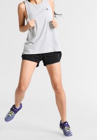 Šedý bezrukávový top s černým logem, doplněný černými šortkami. Fialové sportovní boty mají žluté tkaničky a šedé detaily.