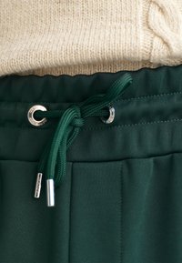 Zielone spodnie na elastycznej talii z zapięciem na sznurek, z metalowymi oczkami w kolorze srebrnym i gładkim, teksturowanym materiałem. Uzupełnione kremowym, dzianinowym topem.