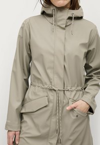 Lichtbeige regenjas met een capuchon, trekkoord in de taille en waterdicht materiaal. Voorzien van twee voorzakken en accenten in zilverkleurig metaal.