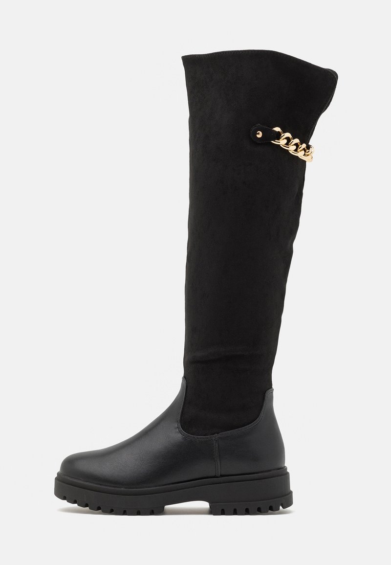 Botte noire haute jusqu'au genou avec semelle épaisse, pied en cuir lisse, tige supérieure en daim et détail décoratif de chaîne en or près du dessus.