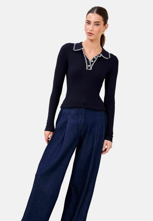 Frau trägt ein eng anliegendes, schwarzes, geripptes Langarmshirt mit weiß abgesetztem Kragen und Knopfleiste, kombiniert mit weiten, dunkelblauen Hosen.