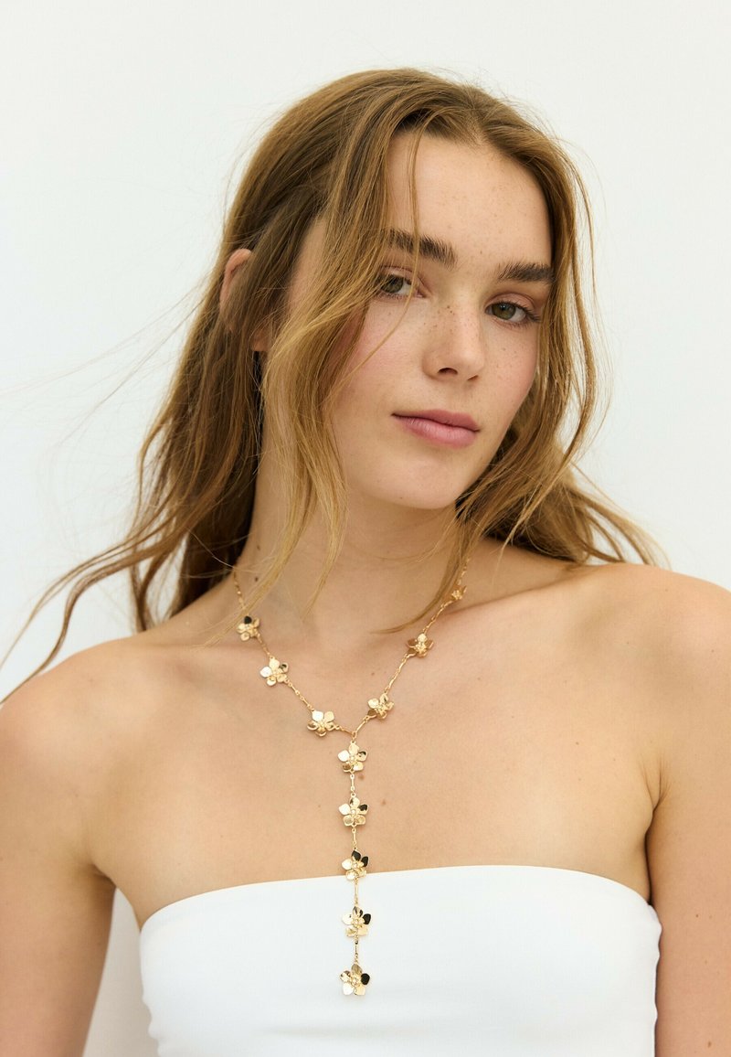 Jeune femme aux cheveux châtain clair portant un top blanc sans bretelles et un collier doré fleuri sur un fond clair uni.