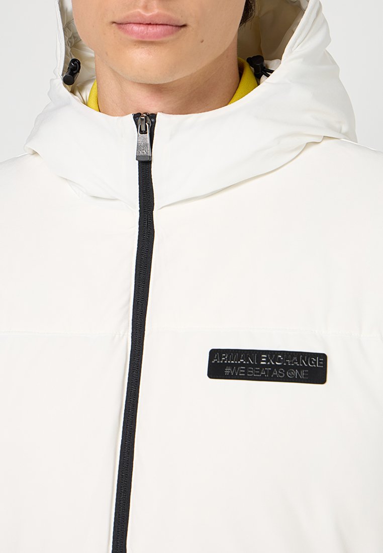 Giacca a vento bianca in materiale imbottito con chiusura a zip frontale, cappuccio regolabile e patch logo nero. Presenta una trama liscia e un design minimalista.