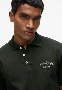 Polo vert foncé en tissu texturé, patte de boutonnage, et logo brodé blanc sur la poitrine, avec un col classique.