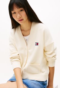 Kremfarget strikket cardigan med V-hals, knappelukking, ribbede mansjetter og en frontlomme. Vist med et logo-patch på venstre bryst.
