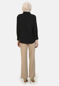 Black long-sleeve blouse with a rounded hem, paired with beige wide-leg trousers and light beige hijab, standing in neutral background.