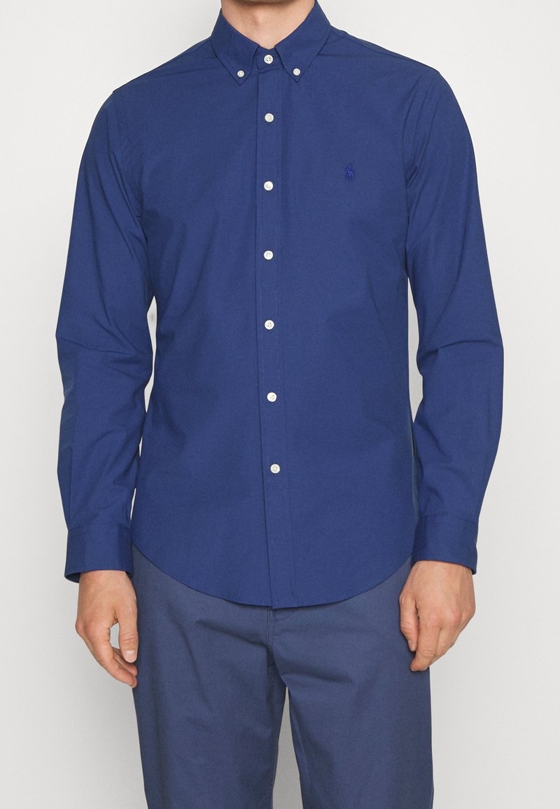 Camicia a maniche lunghe blu navy con colletto a punta e bottoni bianchi. Presenta un piccolo logo sul lato sinistro del petto e orlo dritto.
