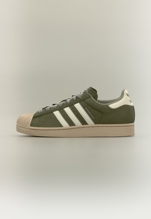 Scarpa da ginnastica verde con strisce bianche, puntale in gomma, design con lacci e suola beige opaca. Presenta il logo del marchio sui lati e sulla linguetta.