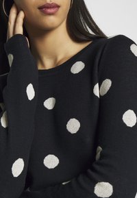 Tummansininen villapaita, jossa on suuria valkoisia pilkkuja. Pehmeä neulottu kangas, pyöreä pääntie ja pitkät hihat. Pilkkujen teksturoitu yksityiskohta.