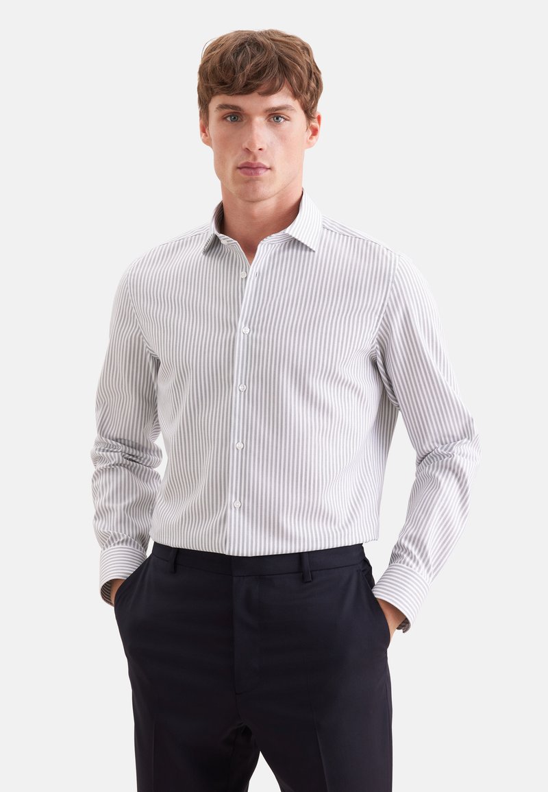 Jeune homme portant une chemise blanche et grise à rayures verticales, rentrée dans un pantalon noir, mains dans les poches, sur fond blanc.