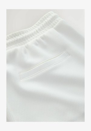 Pantaloni albi din țesătură texturată cu talie elastică și buzunar posterior orizontal cu deschidere, prezentate în prim-plan detaliat.