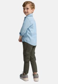 Camisa de botón azul claro, pantalones cargo verde oscuro con bolsillos laterales y zapatillas grises. Niño mostrado de espaldas, sonriendo por encima del hombro.