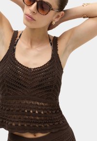 Mujer con gafas de sol carey, pendientes de aro dorados y un top marrón de crochet sin mangas, con las manos detrás de la cabeza.
