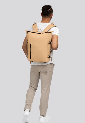 Uomo con uno zaino beige a collo rovesciato con cerniere nere, pantaloni beige, t-shirt bianca e sneakers bianche, in piedi contro uno sfondo grigio.