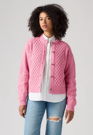 Woman with curly hair wearing a pink knitted cardigan over a white button shirt and ripped light blue jeans standing against a plain background.