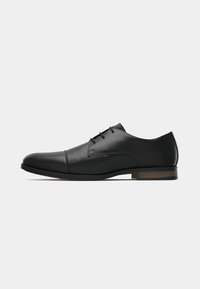 Zapato de vestir oxford para hombre en cuero negro, con punta redondeada, costura en la punta, tres ojetes para los cordones y tacón bajo en color marrón, vista lateral.