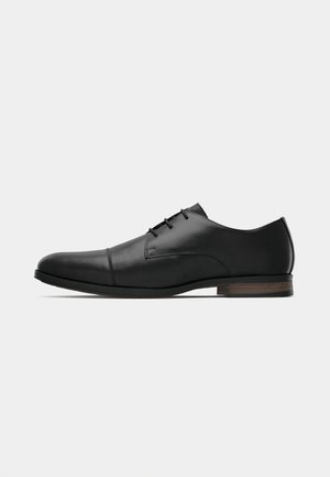 Black leather men's oxford dress shoe with rounded toe, cap toe stitching, three lace eyelets, and low stacked brown heel, side view.