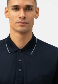 Polo shirt blu navy con colletto a contrasto con bordo bianco, tre bottoni, tessuto testurizzato e un piccolo dettaglio con taschino sul petto.