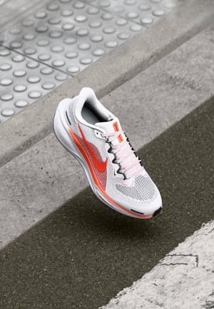 Zapatilla de correr Nike blanca y naranja flotando sobre superficies texturizadas de hormigón y asfalto cerca de un bordillo y pavimento táctil.