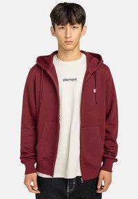 Bordeauxrode zip-up hoodie met een capuchon, twee voorzakken, geribbelde manchetten en zoom, en een klein logo op de linkerkant van de borst, gedragen over een beige t-shirt.