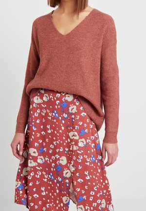 Femme portant un pull à col en V côtelé de couleur rouille, rentré dans une jupe midi rouge avec un motif floral abstrait bleu, crème et rose.