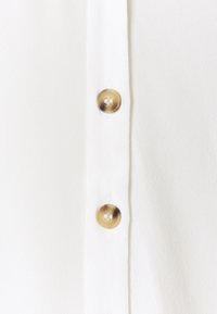 Camisa blanca con un cierre frontal de botones y dos botones redondos de color marrón claro. La tela tiene una textura sutil.