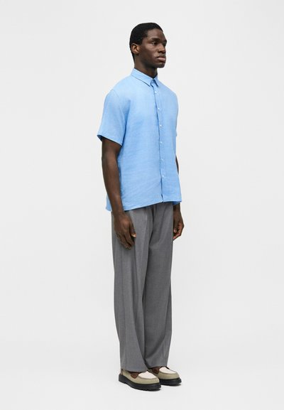 Άνδρας που στέκεται φορώντας ένα ανοιχτό γαλάζιο κοντομάνικο πουκάμισο με κουμπιά, γκρι παντελόνι με φαρδιά μπατζάκια και δίχρωμα loafers σε μονόχρωμο φόντο.