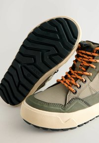 Hoge sneakers met een bovenwerk van khaki stof met textuuraccenten, oranje veters en een robuuste zwarte rubberen zool met een zigzagpatroon.