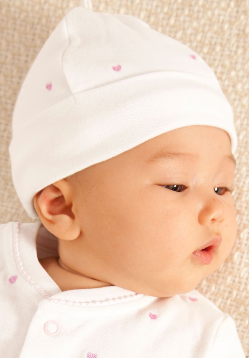 Cappellino bianco per neonati con motivi a cuore rosa; tessuto morbido, vestibilità aderente. Body bianco con dettagli rosa e piccoli bottoni, sfondo semplice.