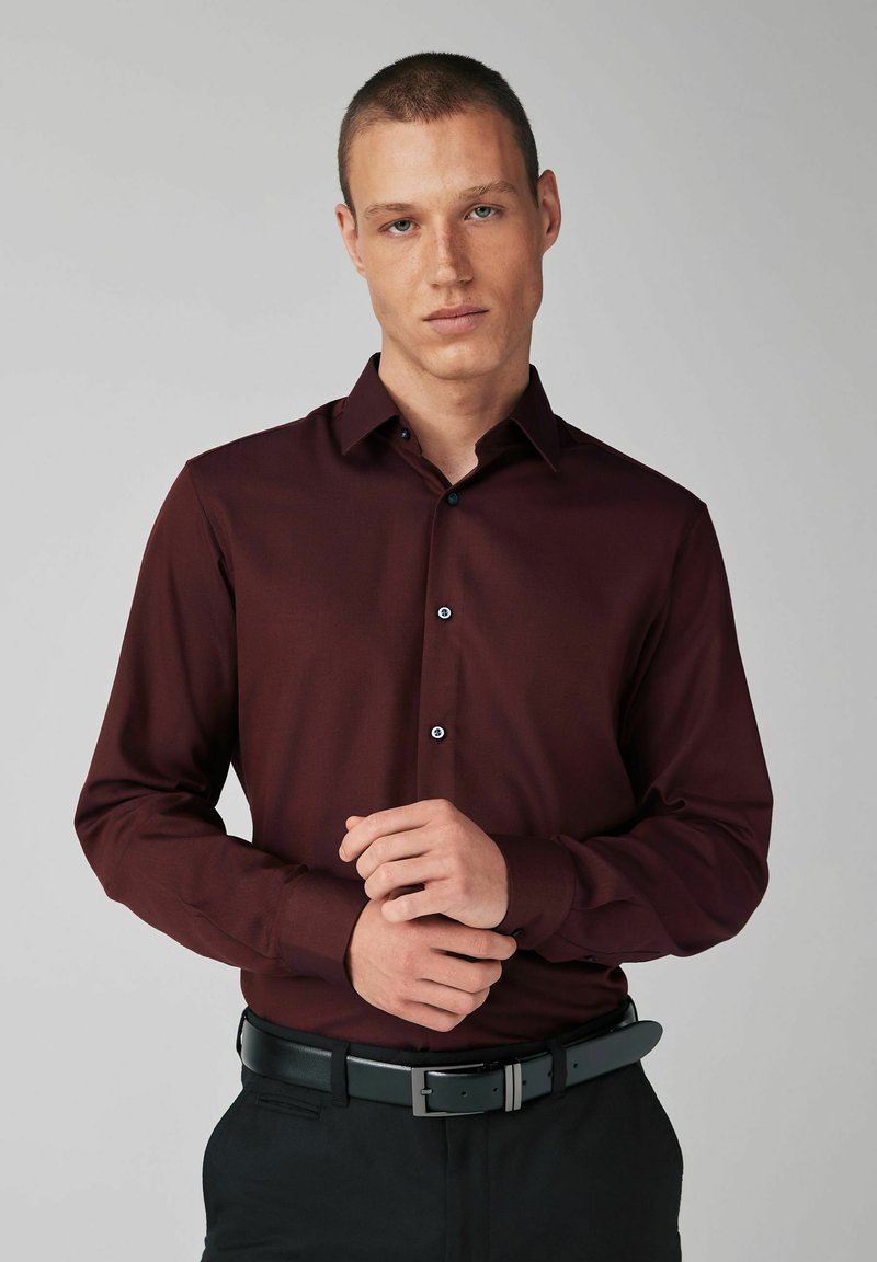 Burgunderfarbene Hemdbluse mit langen Ärmeln, Knopfleiste, schwarzen Knöpfen und glatter Stofftextur, getragen mit einem schwarzen Gürtel und dunklen Hosen.