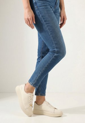 Frau trägt blaue Skinny-Jeans und cremeweiße Plateau-Sneaker, steht mit einem leicht angewinkelten Bein.