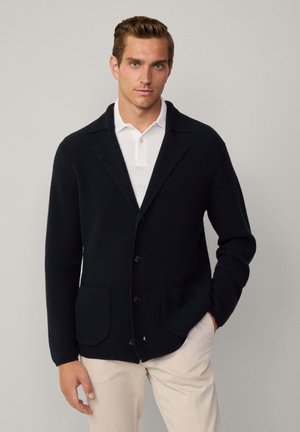 Zwarte gebreide cardigan met een gestructureerd patroon, schouderkraag, enkelvoudige knopen en twee voorzakken. Draagt over een witte polo.