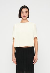 Femme aux cheveux courts et foncés portant un chemisier blanc transparent à manches courtes et une jupe noire plissée taille haute, debout devant un fond uni.