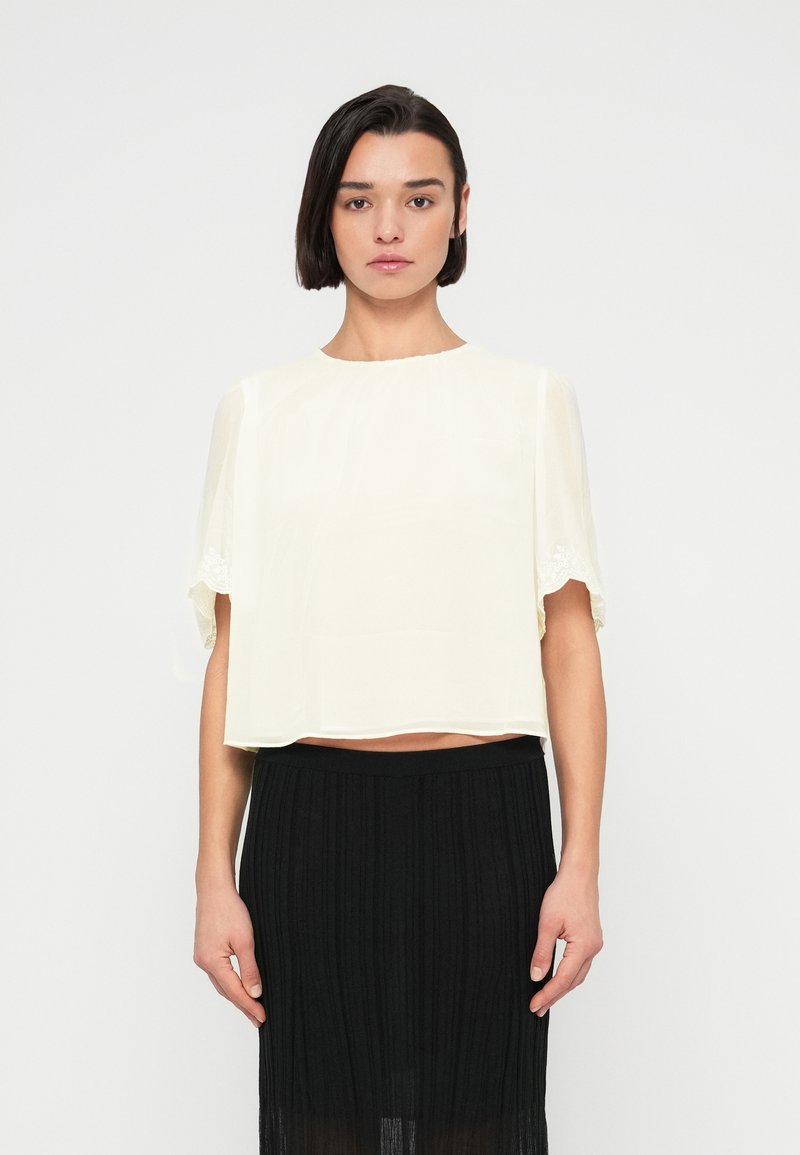 Femme aux cheveux courts et foncés portant un chemisier blanc transparent à manches courtes et une jupe noire plissée taille haute, debout devant un fond uni.