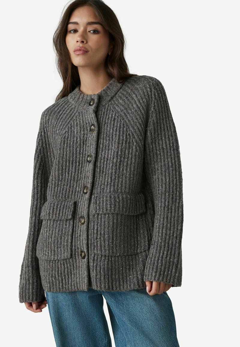 Cardigan gris de canalé con cuello redondo, botones frontales, dos bolsillos tipo parche y tejido texturizado, combinado con jeans azules.
