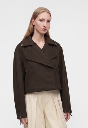 Femme aux longs cheveux blonds porte une veste courte marron foncé avec de larges revers et un pantalon plissé crème taille haute.