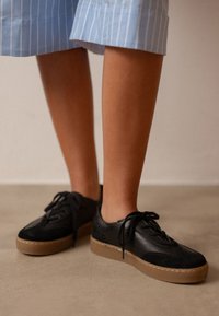 Zapatillas negras con una mezcla de cuero liso y texturizado, suela de goma marrón contrastante y diseño de cordones. Las piernas del modelo son visibles.