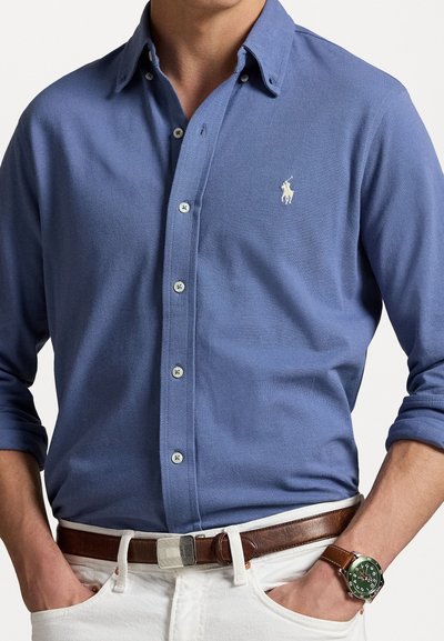 Camicia azzurra a maniche lunghe con tessuto testurizzato e un piccolo logo, indossata con una cintura marrone e un orologio argento e verde, abbinata a pantaloni bianchi.