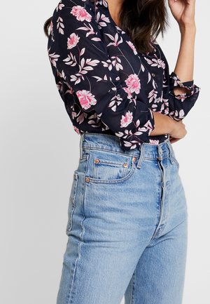 Frau trägt ein schwarzes langärmliges Hemd mit Blumenmuster, in hellblaue, hoch taillierte Jeans gesteckt, mit hochgekrempelten Ärmeln.