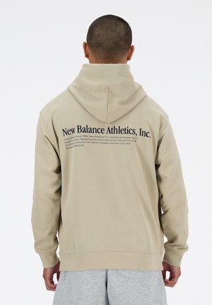 Personne aux cheveux courts portant un sweat à capuche beige avec un texte noir "New Balance Athletics, Inc." au dos, assorti à un pantalon de survêtement gris.