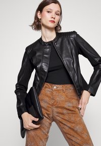 Jaqueta cropped de couro preta com ombros em gomos, combinada com calças marrons texturizadas que apresentam um padrão de manchas e uma clutch preta.