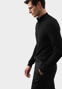 Černý svetr na zip vyrobený z hladké, lehké tkaniny. Přiléhavý střih, žebrované manžety, s bočním pohledem na modelku, která ho nosí.