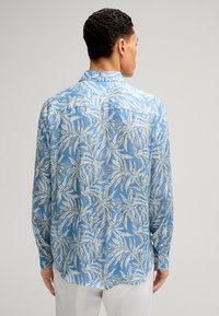 Lichtblauw overhemd met lange mouwen, voorzien van een bladpatroon in wit en groen. Het heeft een button-down kraag en knoopsluitingen bij de mouwen.