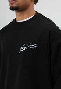 Zwart sweatshirt van katoen met een ronde hals. Voorzien van een voorzak en witte geborduurde tekst op de zak. Zachte textuur.