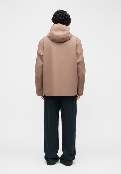 Persona con capelli ricci vista di spalle che indossa una giacca con cappuccio marrone chiaro e pantaloni larghi scuri su uno sfondo uniforme.