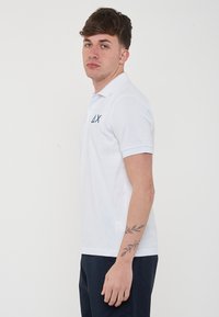 Polo bianco con maniche corte, colletto e logo blu navy sul lato sinistro del petto. Modello che indossa pantaloni blu navy, in piedi di profilo.